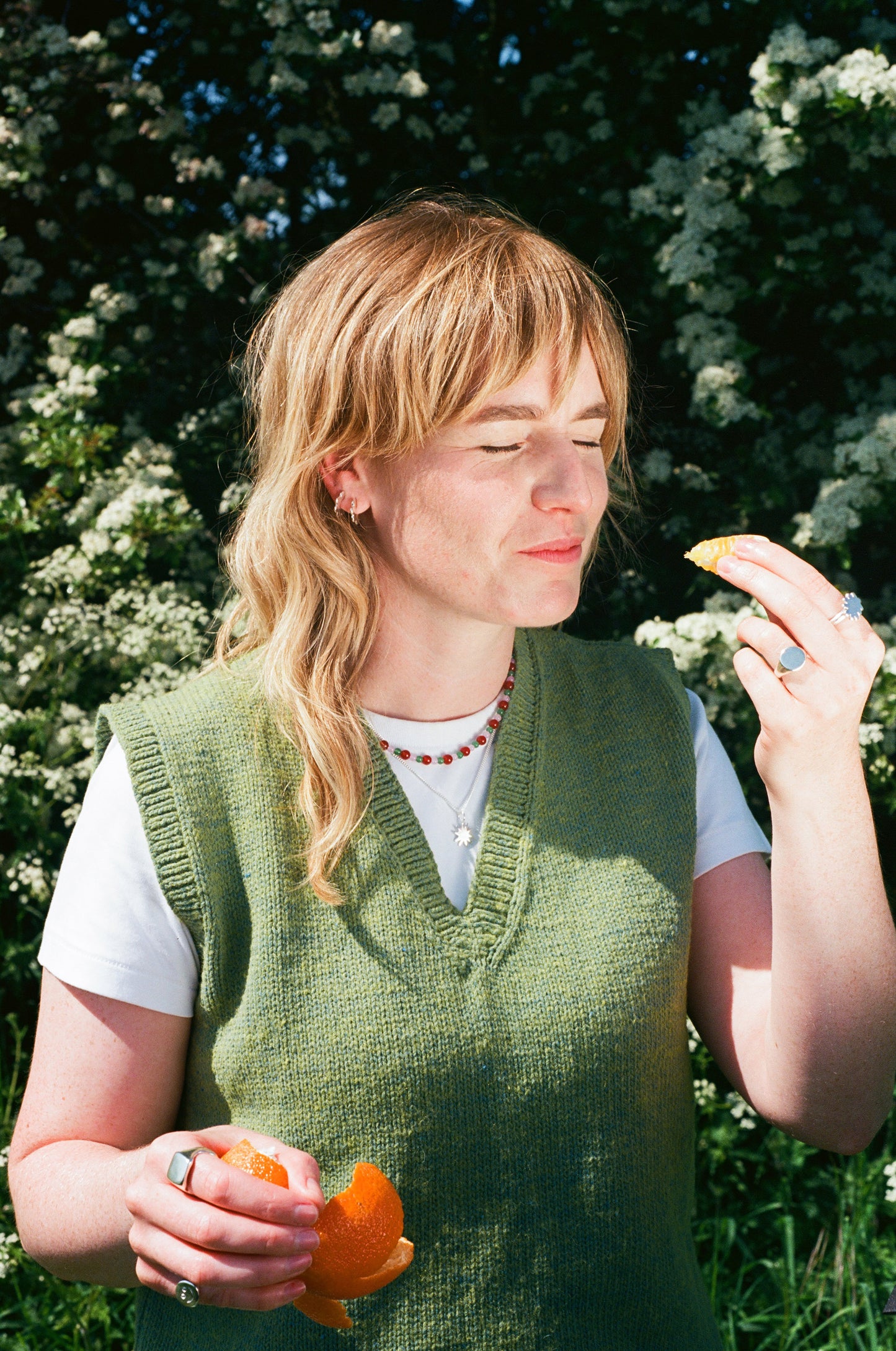 woman smiling eating a tangerine in the sunshine wearing elecectic jewellery in fun playful shapes. scallop hoop earrimgs, flower ring, round signet and beaded necklace. solid recycled silver.