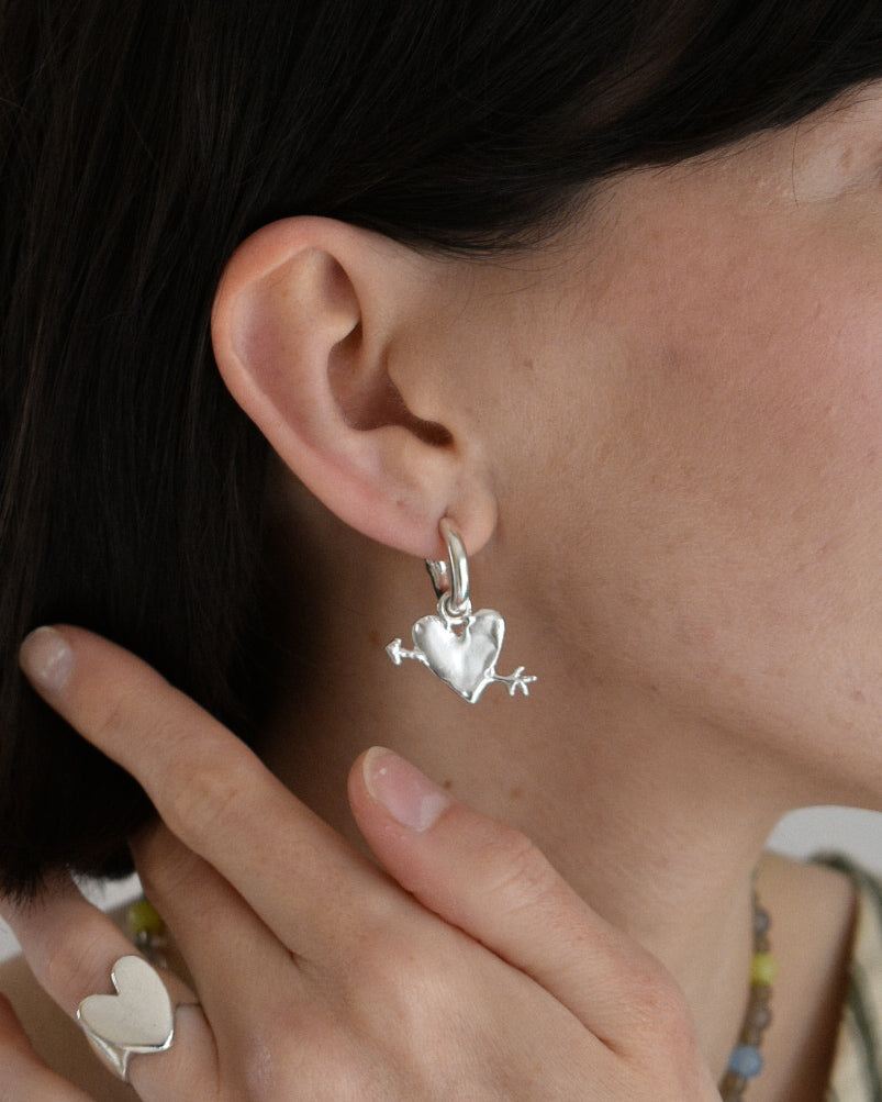 a close up image of a womans ear wearing a hoop earring in sollid recycled silver. the hoop has a heart and arrow charm, with a shiny and wobbly texture