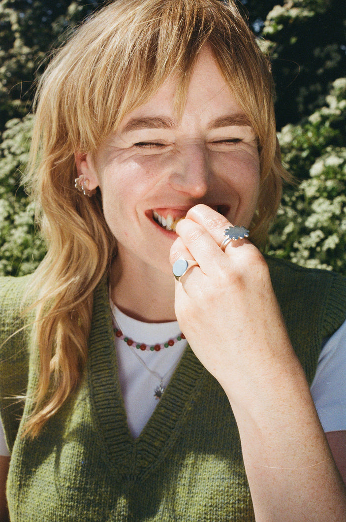 woman smiling eating a tangerine in the sunshine wearing elecectic jewellery in fun playful shapes. scallop hoop earrimgs, flower ring, round signet and beaded necklace. solid recycled silver.