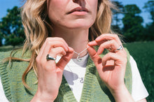 Load image into Gallery viewer, a woman holding her solid silver necklace chain with a bespoke recycled silver initial pendant. she is wearing two solid recycled silver frisbee signets, one plain nd one with a smiley face engraving
