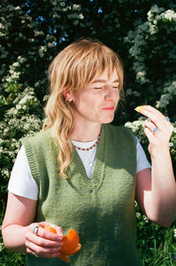 woman wears a dahlia star shaped solid eco recycled silver pendant on a slim silver chain layered with beaded necklace and wearing a green sweater vest. she is holding an orange and wearing silver signet rings.