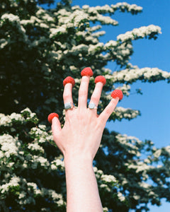 a hand held in the air with raspberries on each finger. the hand wears solid recycled silver signet rings.