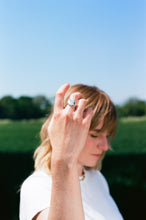 Load image into Gallery viewer, a woman holding up her hand showing a recycled silver shining heart ring with wobbly band. the woman and the field are out of focus with a spotlight on the jewellery.

