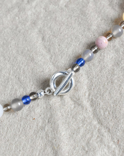 a colourful beaded necklace in a neutral brown grey base with coloirful stone and glass beads scattered. blue jade, agate, carnelian, aventurine. finished with solid recycled silver doughnut toggle clasp.