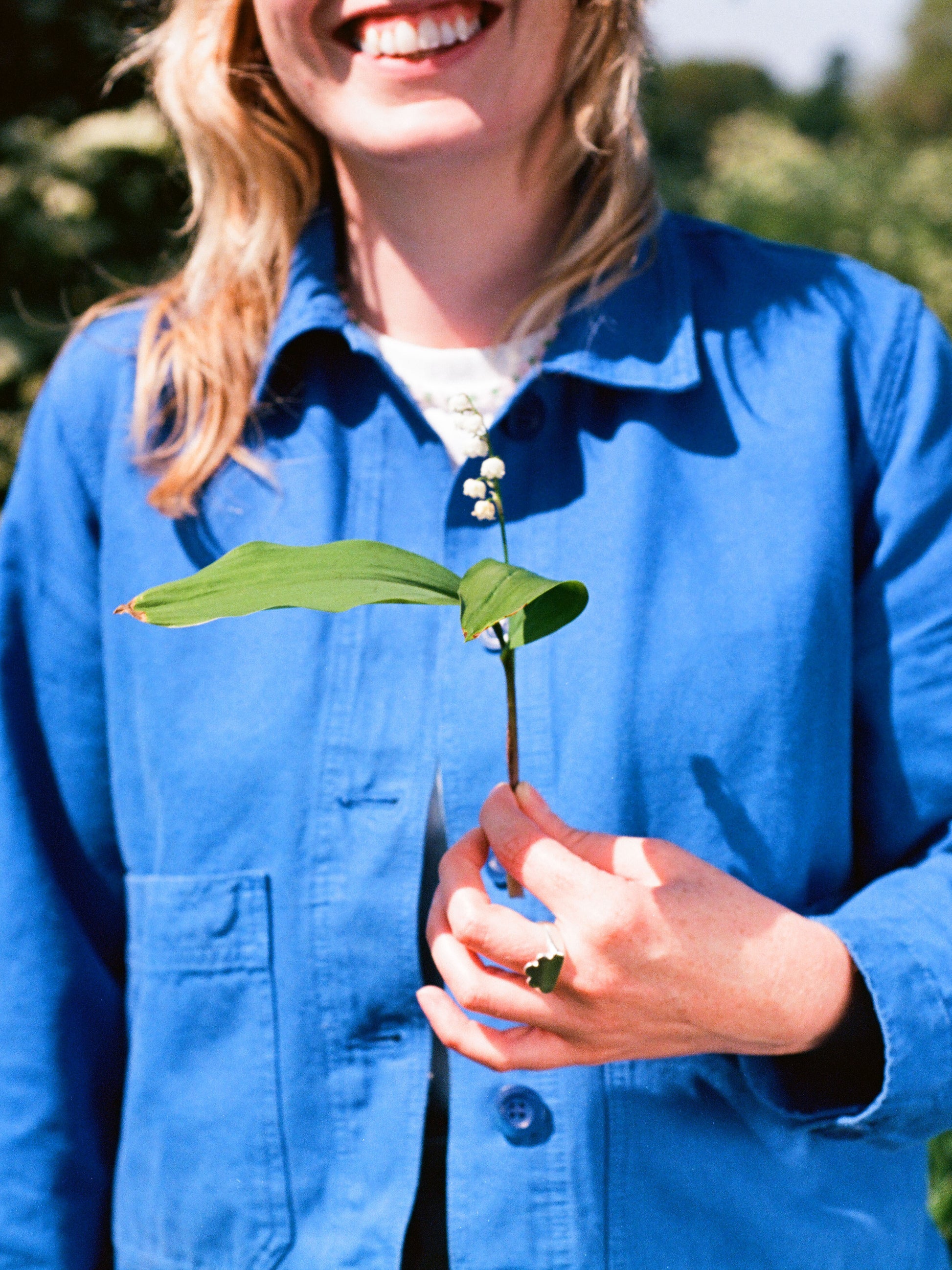 ruth smiling in the summer sunshine  in a royal blue paynter jacket holds a lily of the valley flower and wears a shining solid recycled silver signet ring in the shape of a cloud