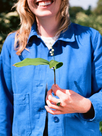 ruth smiling in the summer sunshine  in a royal blue paynter jacket holds a lily of the valley flower and wears a shining solid recycled silver signet ring in the shape of a cloud